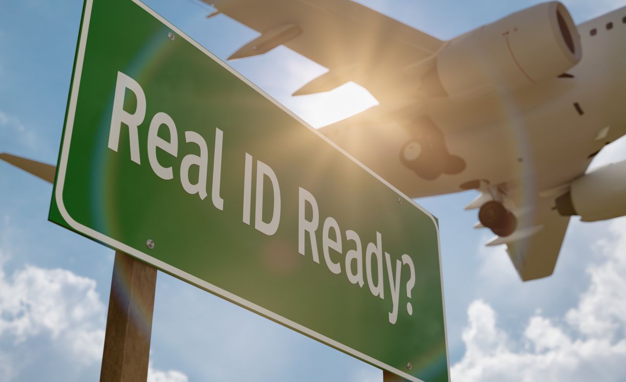 Green “Real ID Ready?” sign with airplane flying overhead, symbolizing federal REAL ID requirements for domestic air travel.